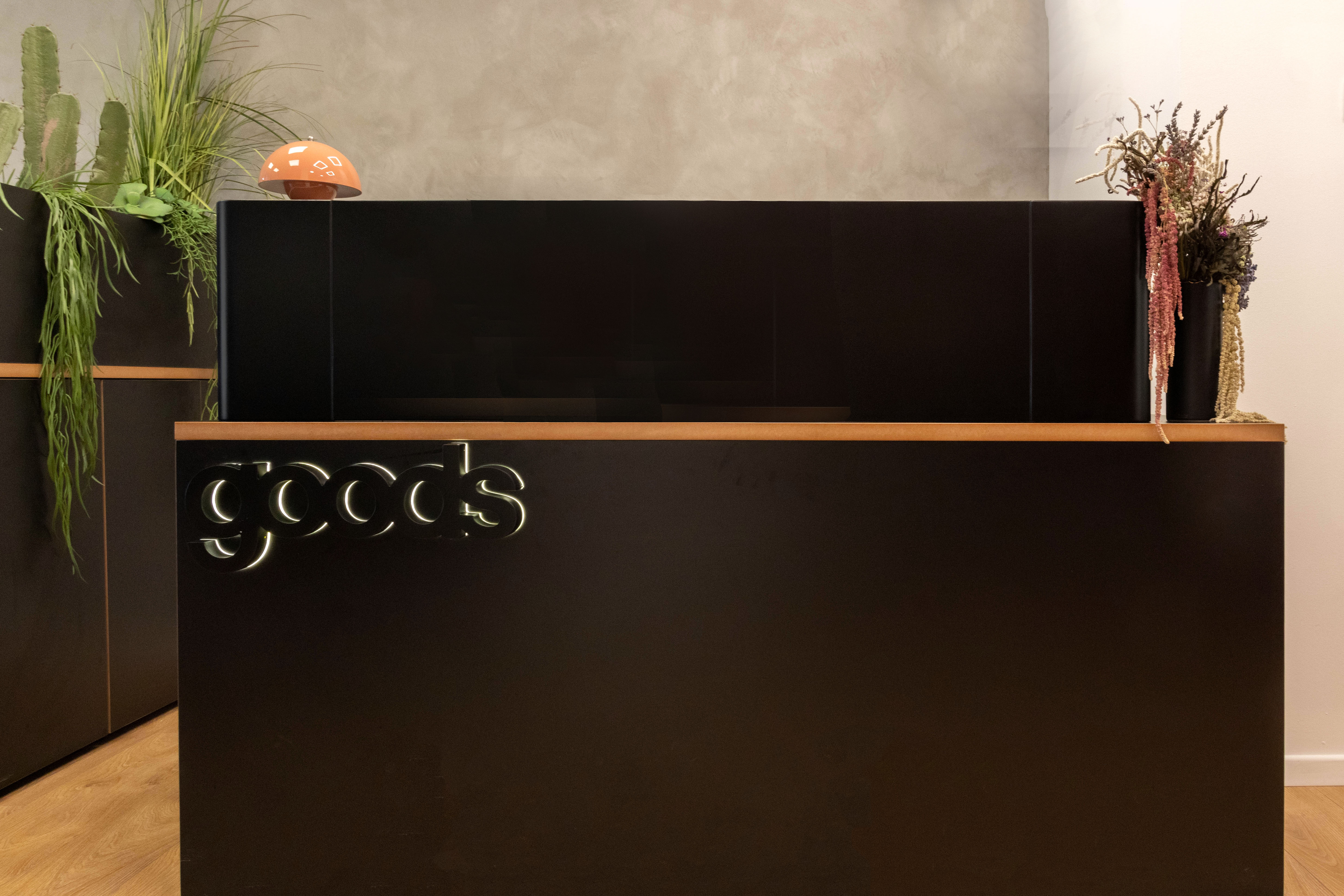 דלפק קבלה שחור עם לוגו 'goods' מואר, עיצוב מינימליסטי עם עציצים יבשים בצדדים, קקטוס בצד שמאל, וקיר טקסטורלי בגוון אפור־ורוד ברקע.